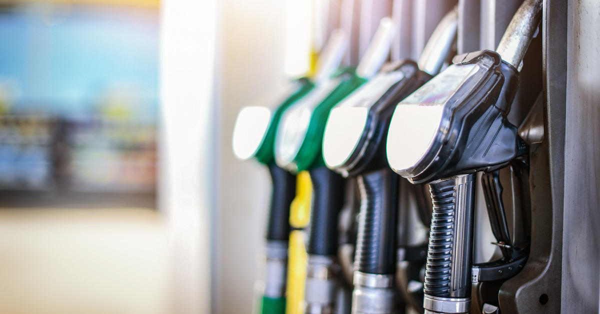 Four different gas pumps, two green and two black, stand out against a blurry gas station and gas pump in the background.