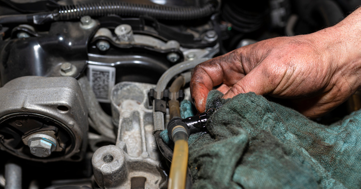 A person with dirty hands cleans off a fuel pump inside the hood of a car with an even dirtier blue rag.