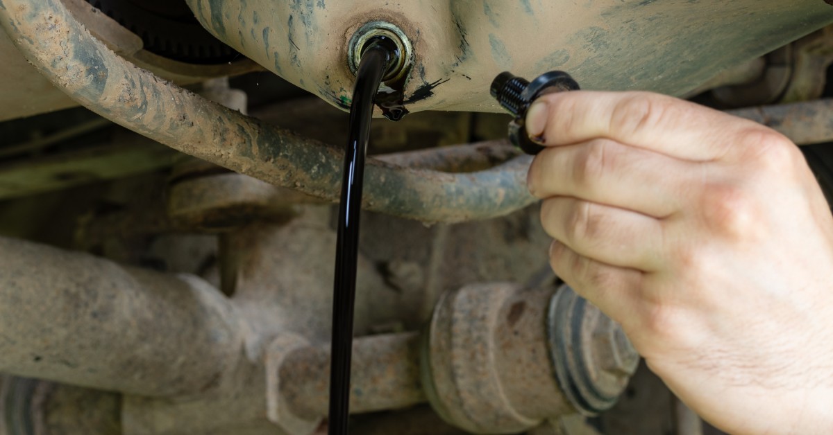 A person holding the drain plug of an oil tank to drain the oil. The oil tank is dirty and it pours out a stream of oil.