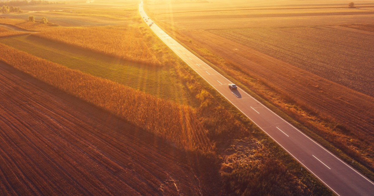 An aerial view of a car driving down an empty road in the countryside with the sun shining over everything.