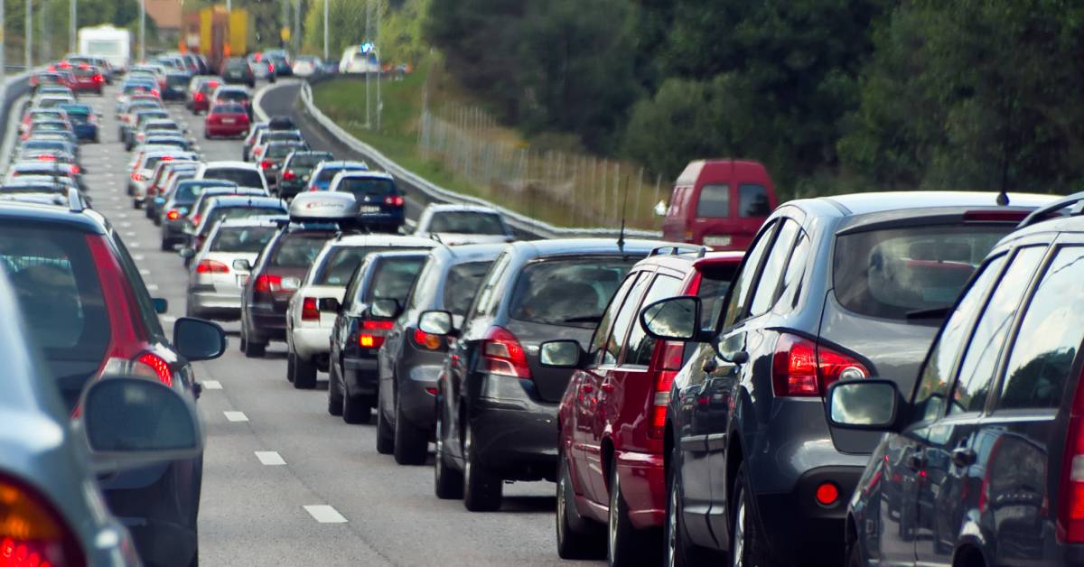 A lot of cars sits in traffic on a freeway. The road curves around some trees, and the traffic is seemingly endless.