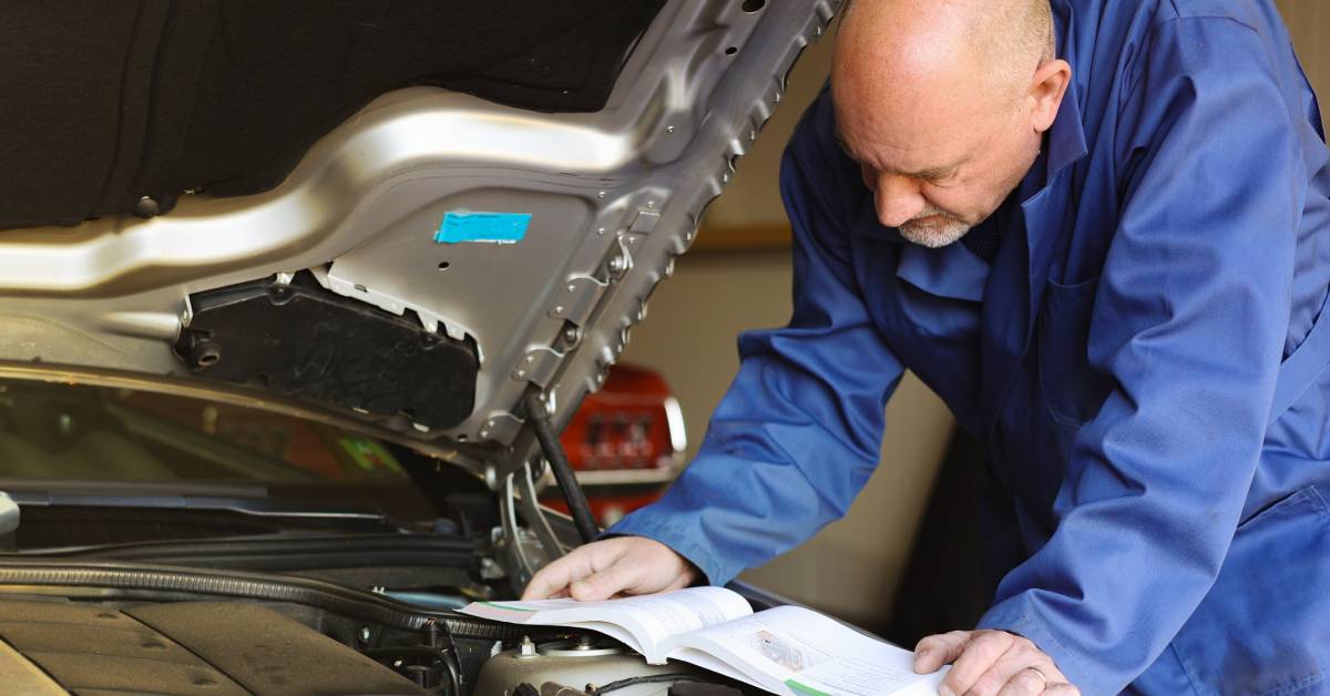 A bald mechanic wearing a blue work suit looks over an open vehicle manual on the open hood of a car.