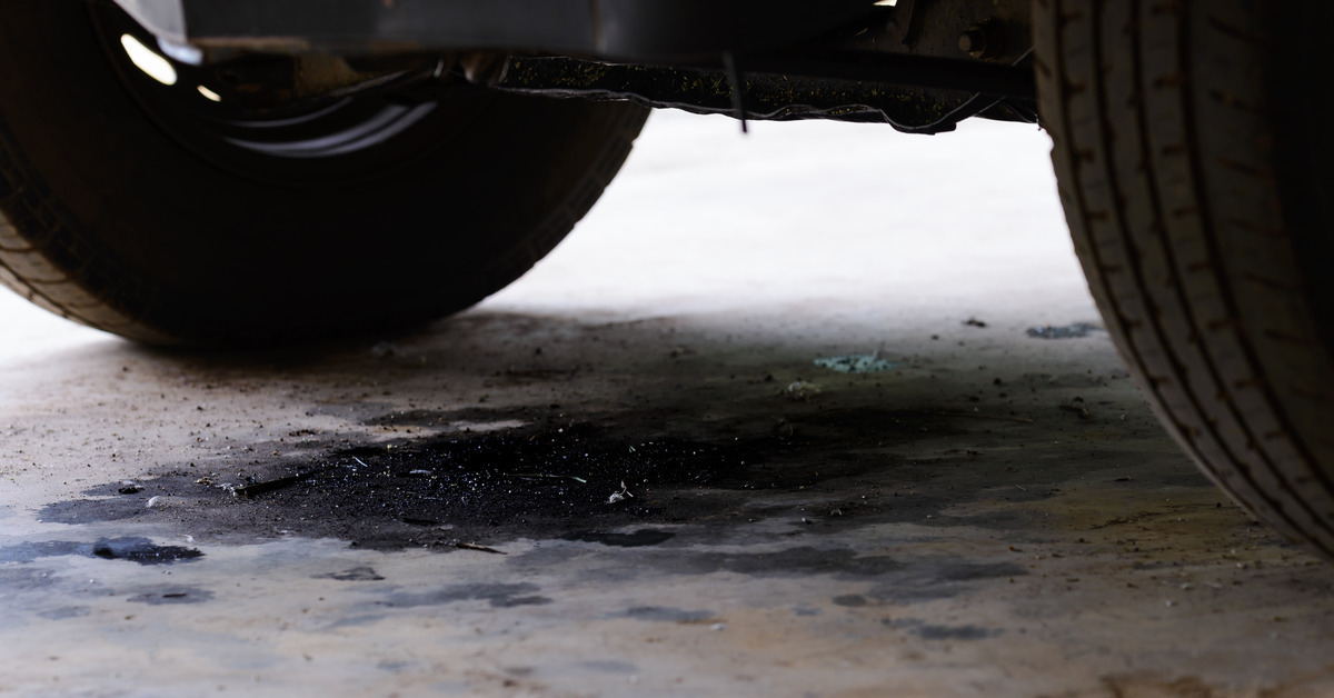 A close-up of an oil leak from a car. There are two tires, and between them, there is a large oil stain on the concrete.