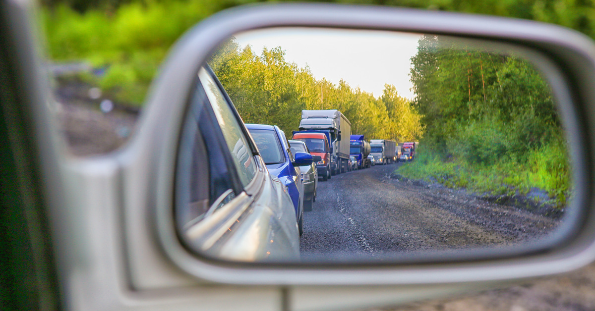 In the view of a car's side mirror, there are multiple cars and semi-trucks idling on a road with lots of trees on the side.