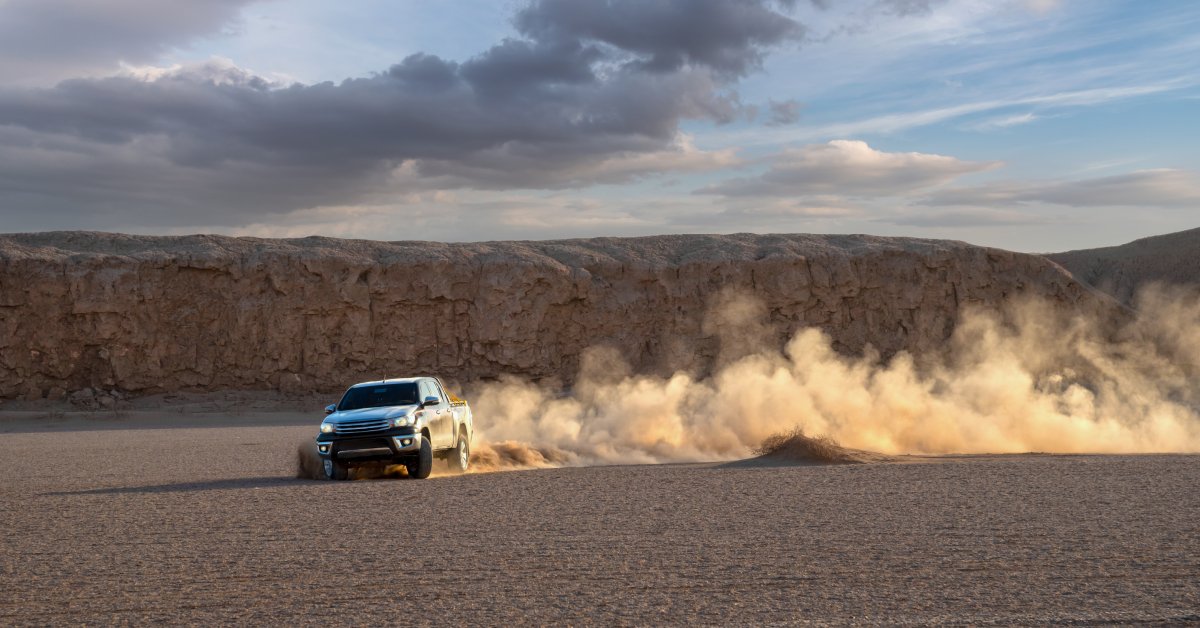 A pickup truck drives across a field of sand and leans to the right while kicking up lots of dust in its wake.