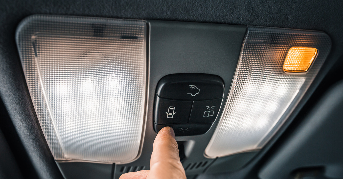 A finger reaches up to press a button next to the interior car dome light on the ceiling. Dark fabric surrounds the light.