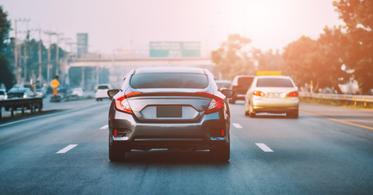 A dark gray car drives down a highway towards the sun in the distance. There are other car on the road.