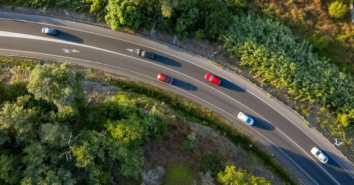 An aerial view of different cars driving down a curved road with green forests on both sides of the road.