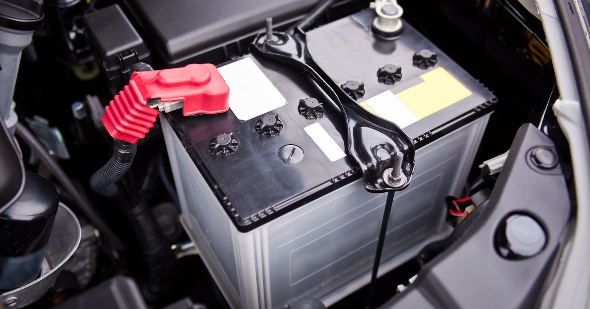 A close up view of a car battery with a red clamp on one part, a large black clamp on another. The battery is in the car.