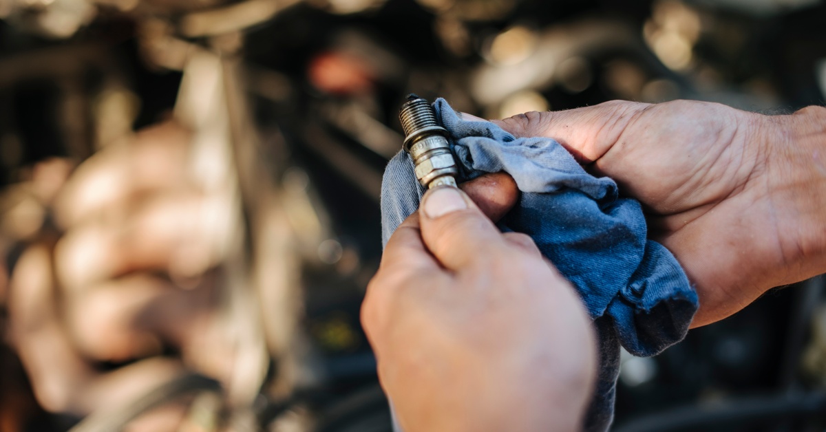 A worker is using a blue cloth to clean a spark plug while the background is a blurry view of a car hood.