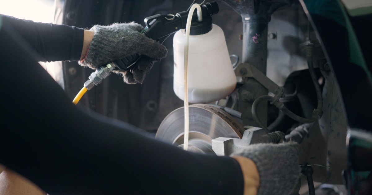 Gloved hands are using a fluid bottle and hose beside an exposed vehicle brake rotor inside a repair workshop.