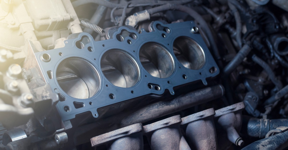 A metal cylinder head gasket being held above an engine block with hoses and circular cylinder openings visible.